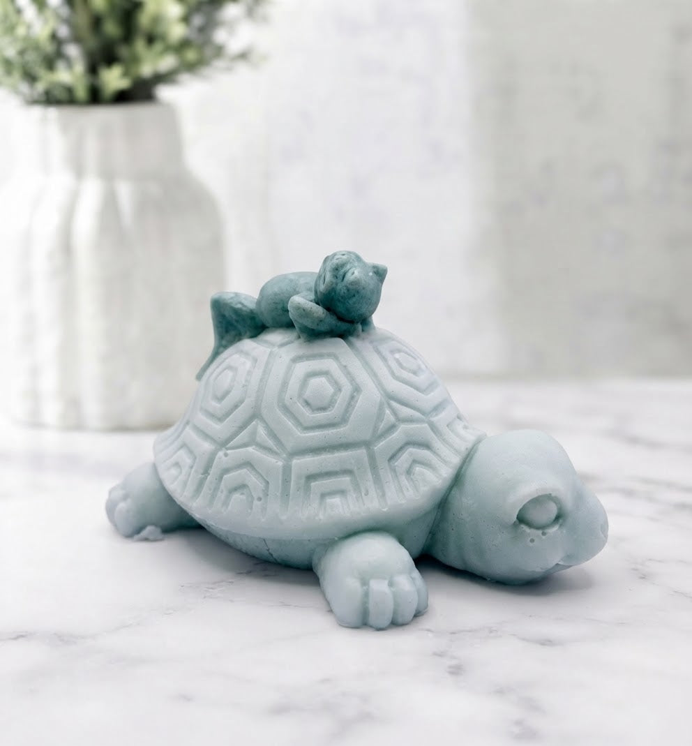 A close-up of a turtle vegan soap with a smaller frog resting on its shell. The turtle faces right on a white marble surface with a blurred floral background 