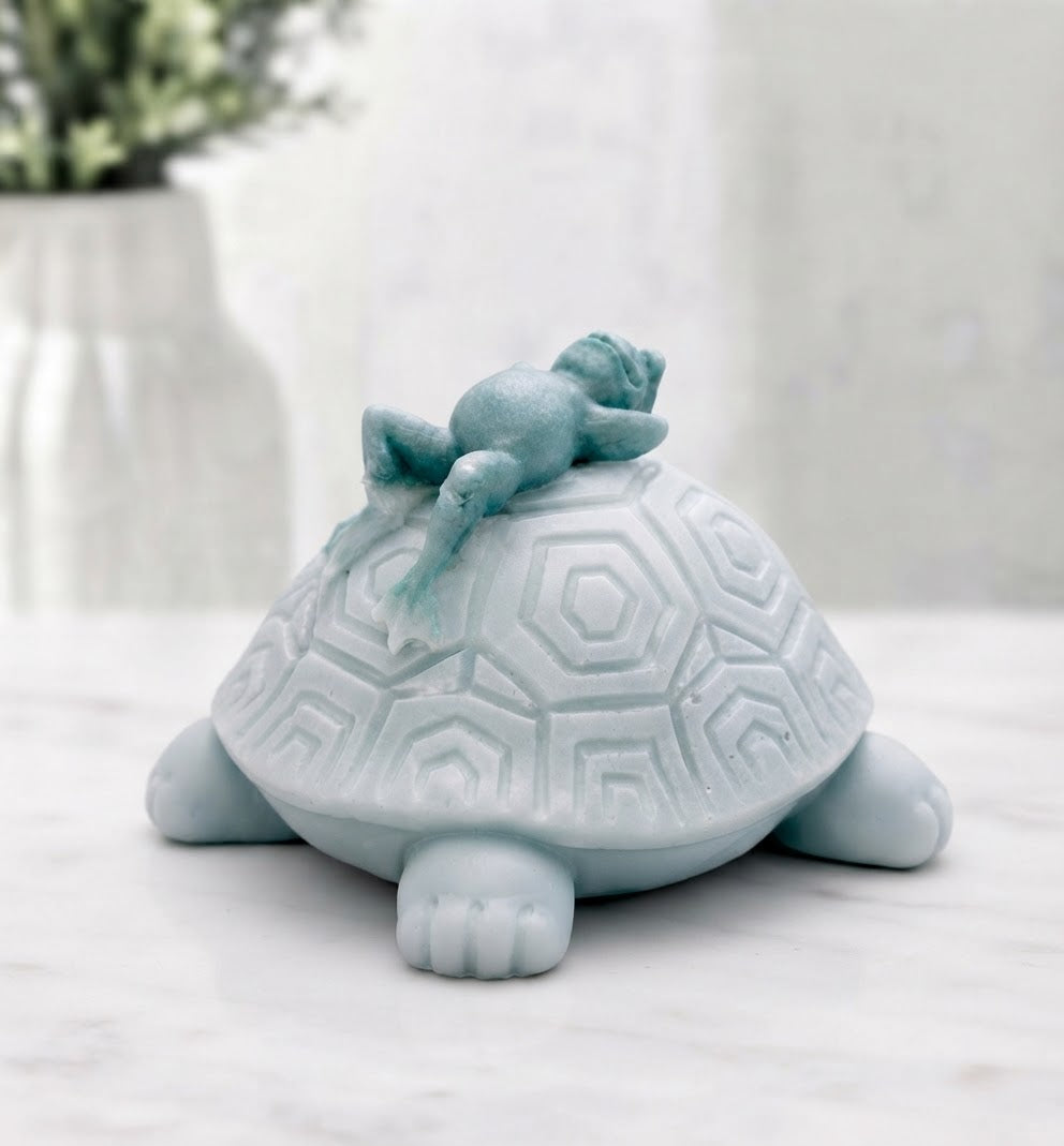 A rear vie of a turtle soap, with a smaller, curled-up frog sleeping on its shell. It rests on a white marble surface with a blurred vase in the background.