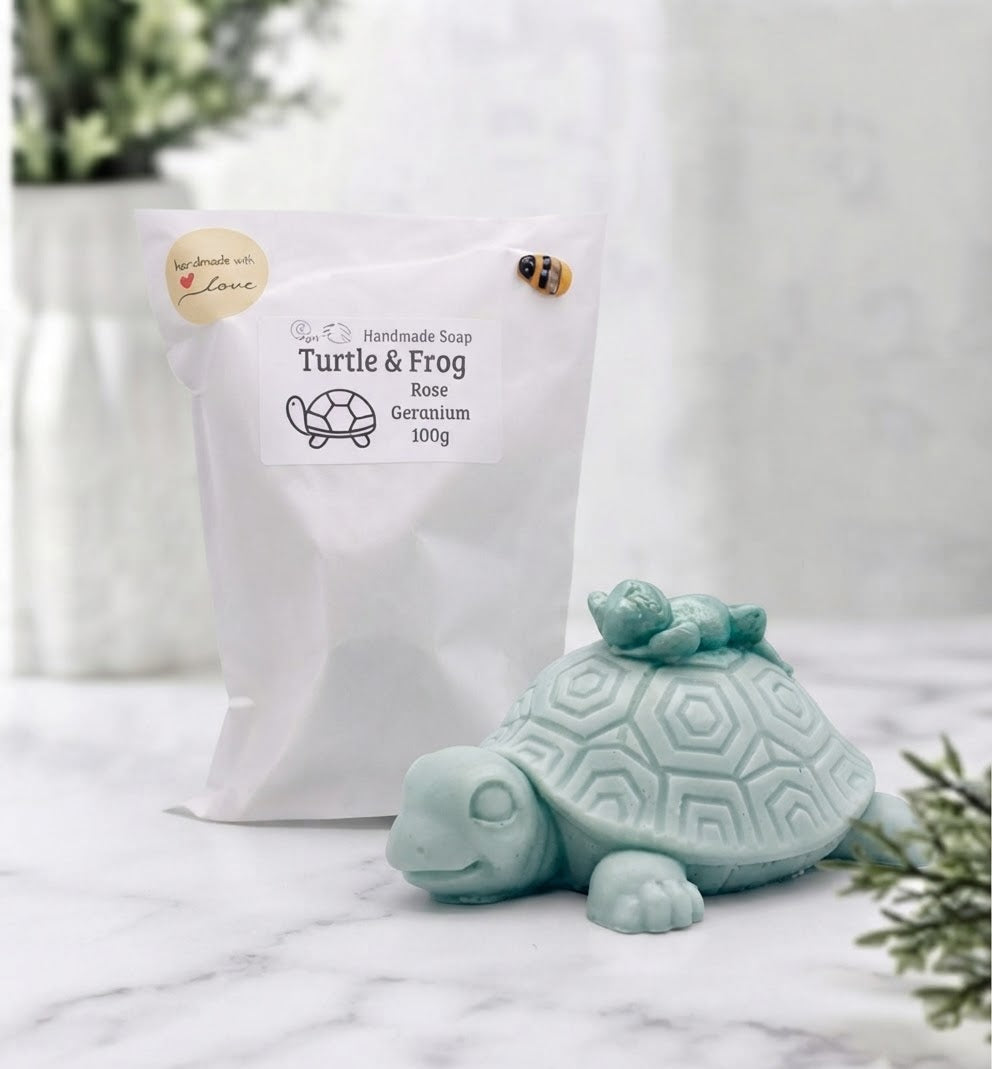 A product shot on white marble showing a white paper soap pouch (with label, heart sticker, and bee) next to a turtle-and-frog soap. Blurred background.