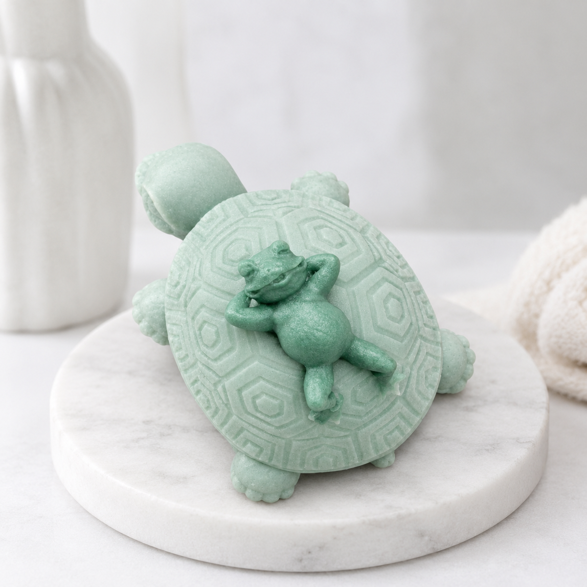 A overhead view of a handmade vegan soap in a soft mint green tone, sculpted into a large friendly turtle with a geometric shell pattern and a small, cute frog sleeping peacefully on its back.