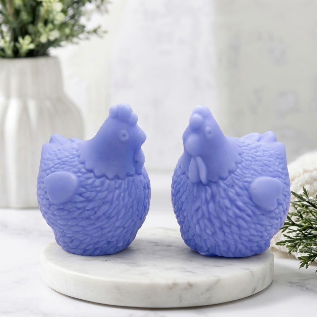 Two soft blue textured chicken-shaped candles sitting side-by-side on a round white marble coaster, with a blurred background of a white vase with greenery.