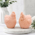 A close-up product photo of a pair of detailed pink chicken-shaped soap bars from the "Little Chickens" gift set, placed on a white marble display stand with a blur background of a flower vase.