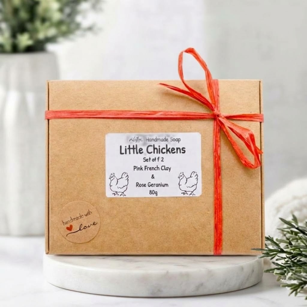 A kraft paper gift box tied with a red ribbon and bow, featuring a white label that reads 'Little Chickens Set of 2 Pink French Clay & Rose Geranium Soaps 80g'. The box is displayed on a white marble pedestal with a soft, blurred background.