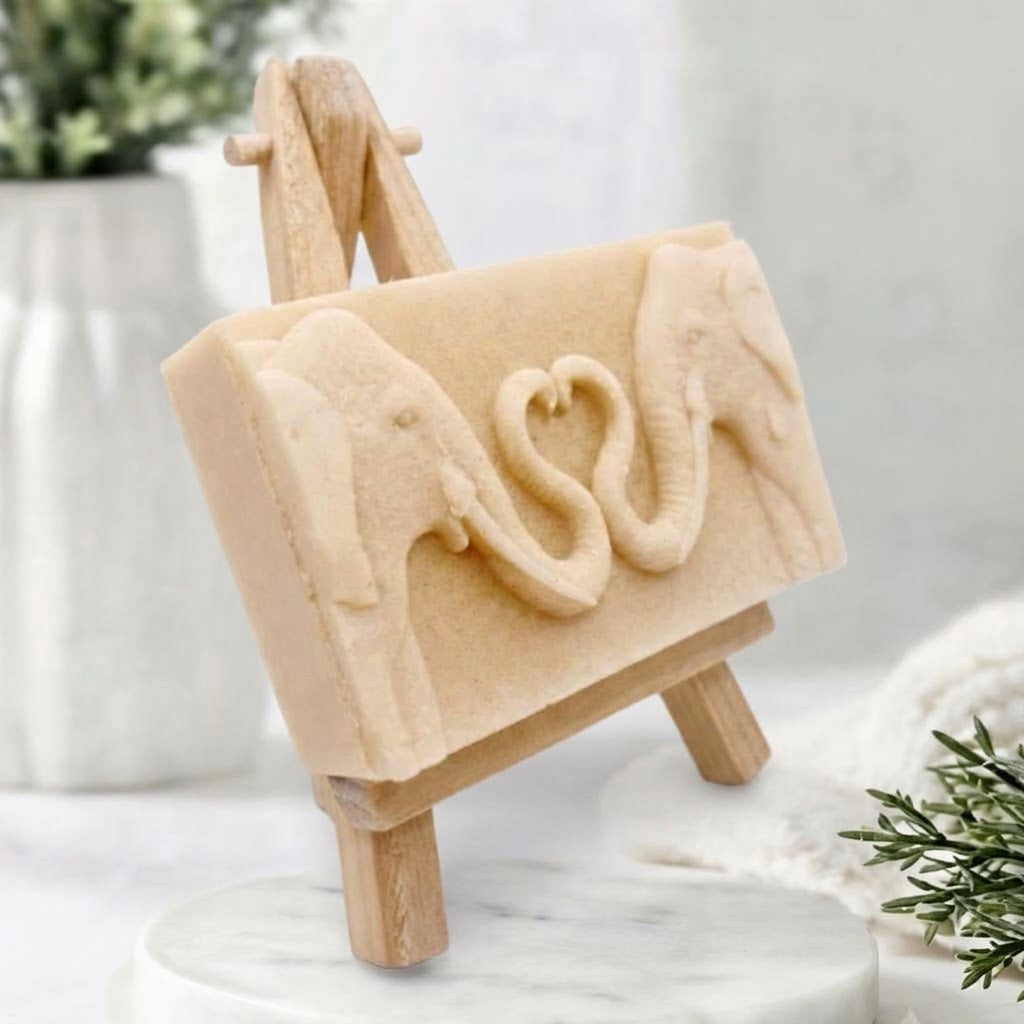 A three-quarter angle view of a cream-coloured artisanal elephant soap bar sitting on a small wooden easel, showing the thickness of the bar and the relief carving of elephants forming a heart on the front.