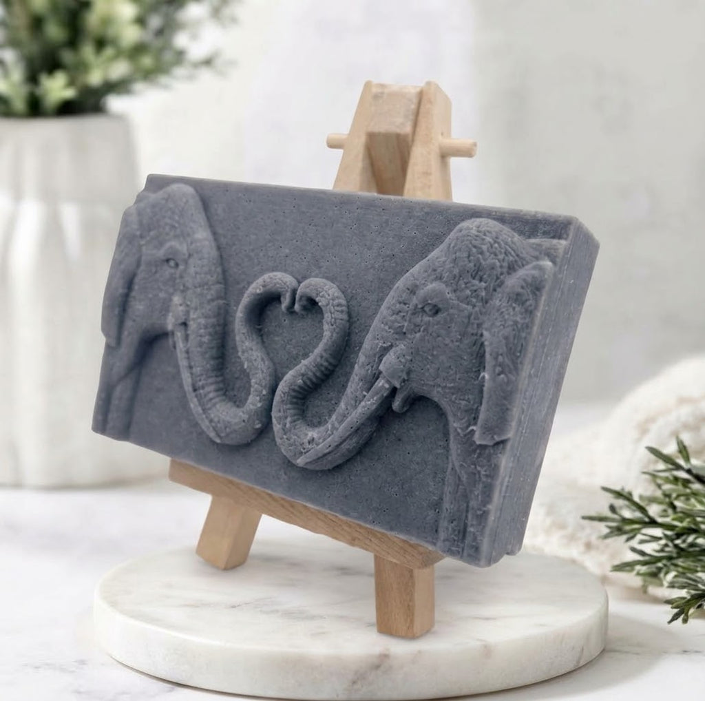 Charcoal grey artisanal elephant soap displayed on a miniature wooden easel. The bar features a sharp relief carving of two elephants with intertwined trunks forming a heart shape.