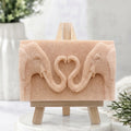 A pink artisanal elephant soap bar displayed on a miniature wooden easel. The bar features a relief carving of two elephants whose intertwined trunks form a heart shape.