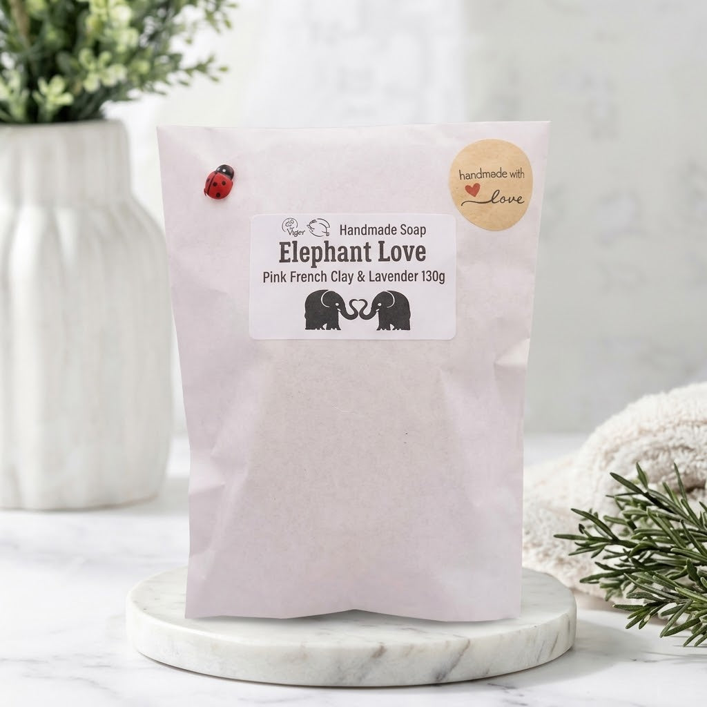 Packaged "Elephant Love" soap in a light pink paper pouch. The pouch is held closed by a miniature red ladybug clip and a "handmade with love" sticker, resting on a marble tray. The main label reads: "Handmade Soap Elephant Love Pink French Clay & Lavender 130g" above a black silhouette of the elephant heart design.