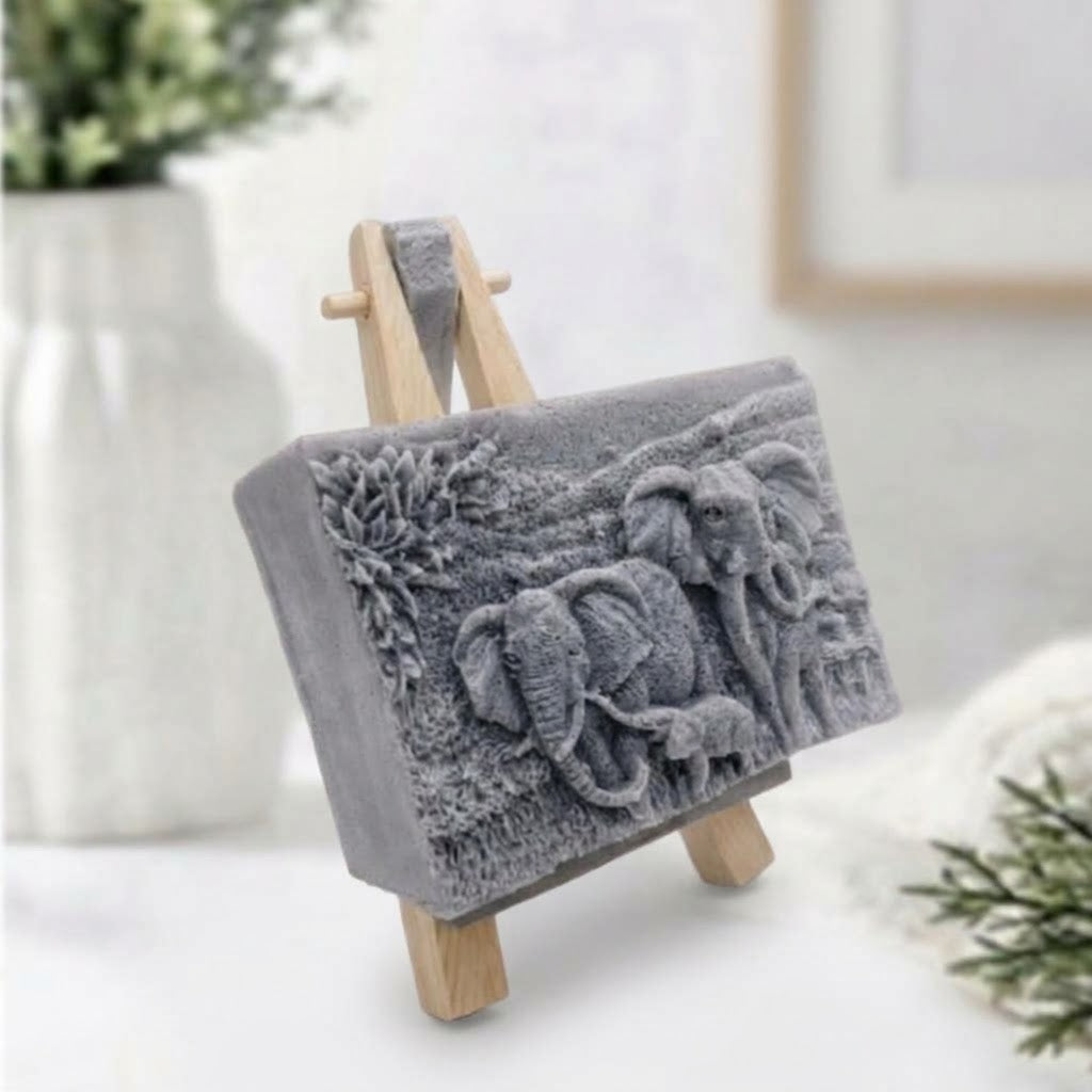 A grey, detailed relief-carved soap slab depicting two larger elephants and one smaller elephant standing amidst carved vegetation, shown at a three-quarter angle resting on a miniature wooden tripod easel.