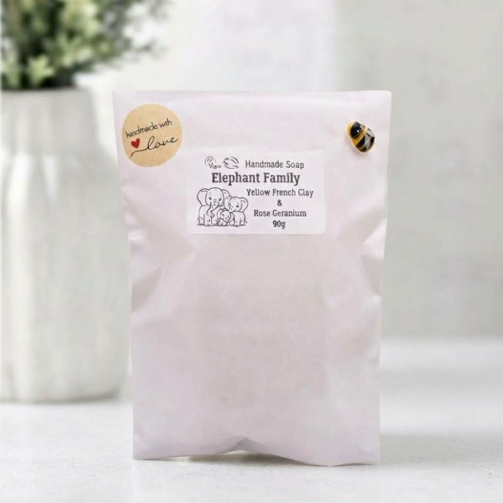 A white paper gift pouch containing an "Elephant Family" handmade soap. The label on the front lists ingredients including yellow French clay and rose geranium, and features a line drawing of an elephant family. The pouch is secured with a "handmade with love" heart sticker on the left and a small yellow and black striped bee on the right.