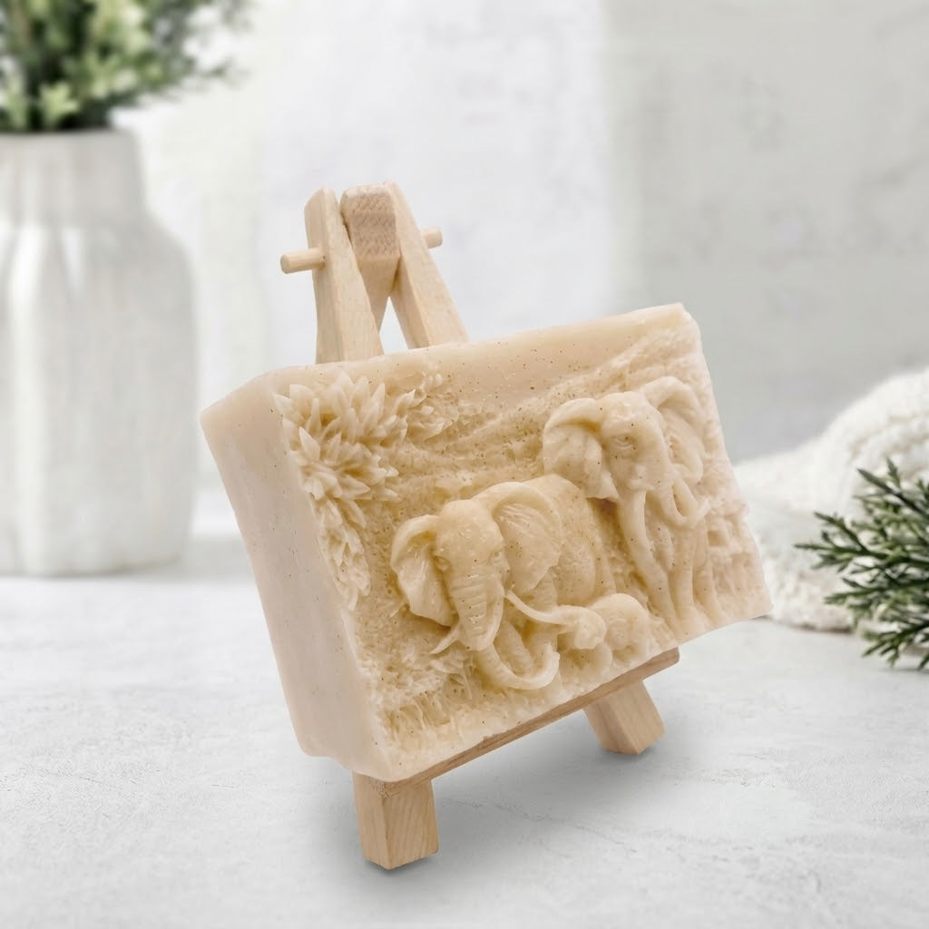 A soft yellow, relief-carved soap slab showing two larger elephants and one smaller elephant amidst carved vegetation, shown from a three-quarter angle supported by a small wooden easel.
