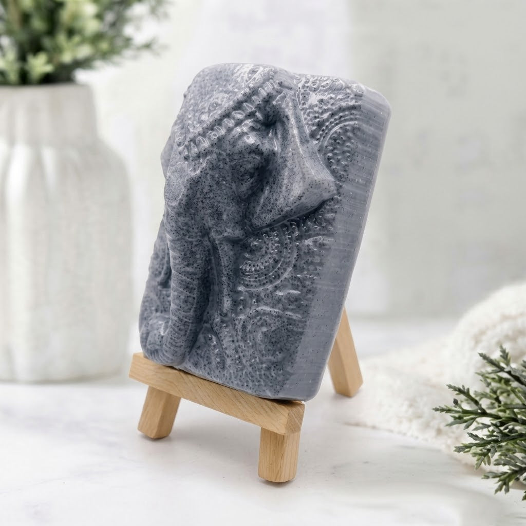 A rectangular bar of grey charcoal soap featuring an intricate high-relief side carving of Ganesha's elephant head and detailed headdress, displayed at an angle on a miniature wooden tripod easel.