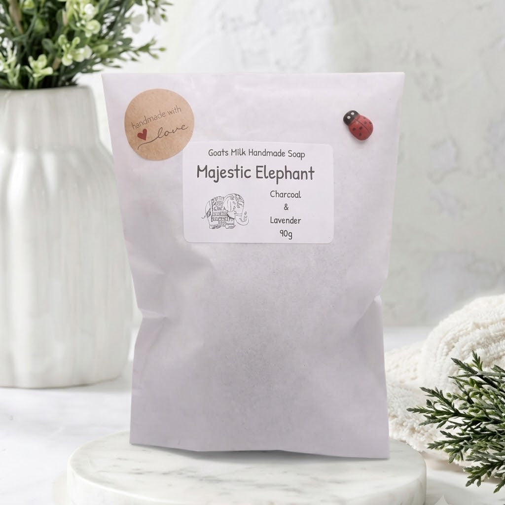 A sealed white paper pouch of Handmade Soap labeled 'Majestic Elephant' with Charcoal & Lavender ingredients and a sketch of an elephant. The pouch is adorned with a 'Handmade with Love' sticker and a small ladybug pin, sitting on a marble stand with a white vase and greenery in the background.