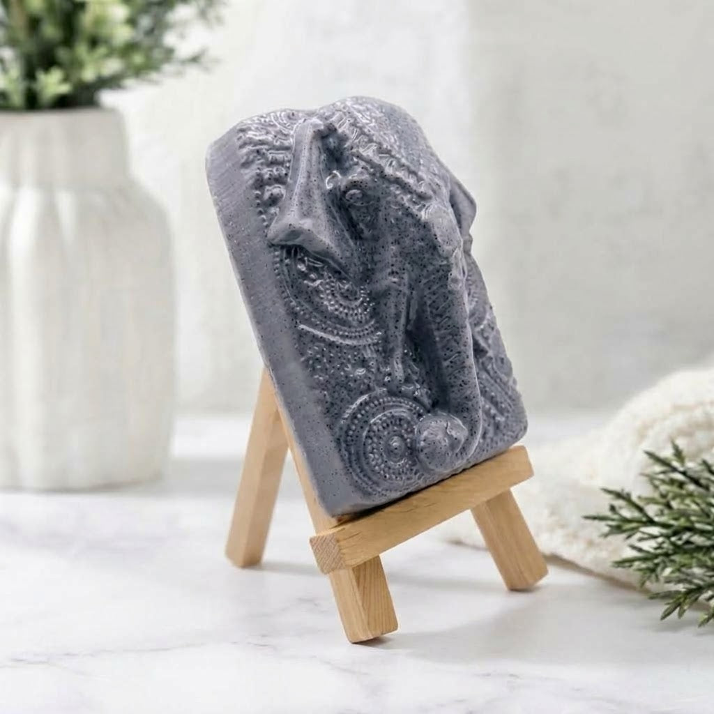 A rectangular bar of grey charcoal soap featuring an intricate high-relief side carving of Ganesha's elephant head and detailed headdress, displayed at an angle on a miniature wooden tripod easel.