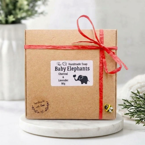 A square, brown kraft paper gift box tied with red raffia is next to two gray, detailed elephant-shaped soap figurines that are sitting on the same patterned bases seen in image_168.png. The box label, "Baby Elephants Charcoal & Lavender,
