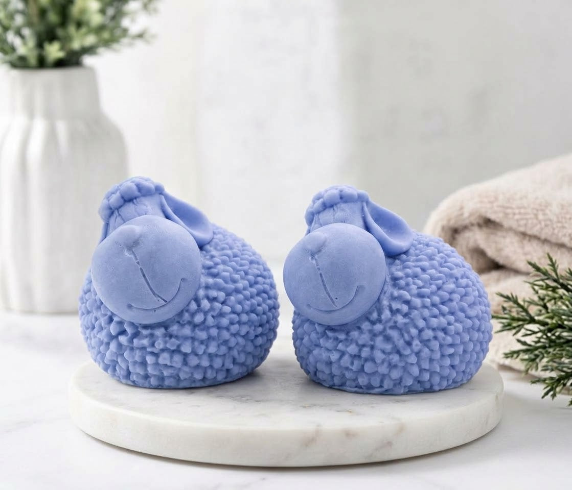 Two cute, pastel blue sheep-shaped soaps with bumpy, textured bodies representing wool, resting on a white marble stand against a background of blurred neutral bathroom decor and towels.