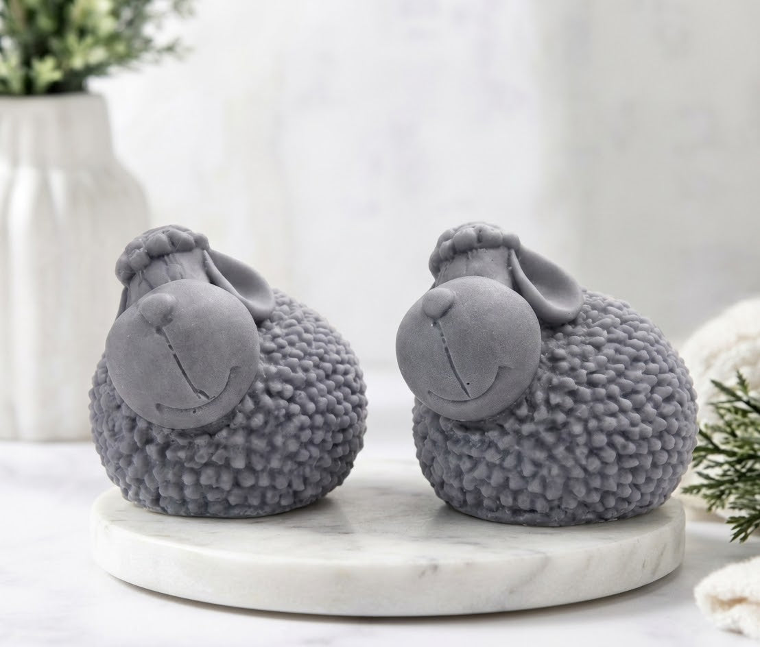 A pair of cute, gray sheep-shaped soaps with highly textured, bumpy wool bodies and smiling cartoon faces, sitting on a round white marble trivet in a blurred bathroom setting.