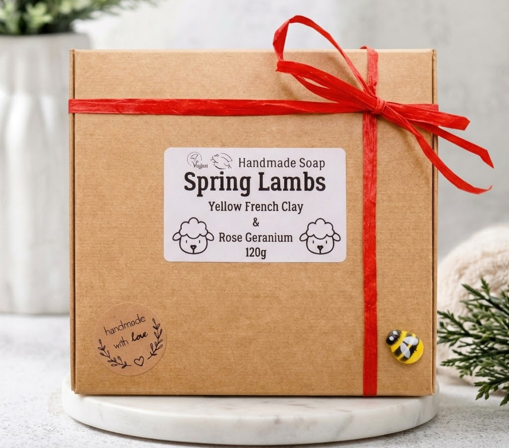 A brown kraft paper gift box for the "Spring Lambs" handmade soap set, tied with a red raffia ribbon and decorated with a "handmade with love" stamp and a small ceramic bee charm. The central white label features line drawings of sheep and lists the scent as "Yellow French Clay & Rose Geranium 120g."