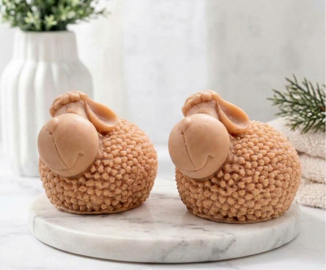 Two cute, terracotta sheep-shaped soaps with smiling faces and detailed, bumpy wool textures, resting on a white marble trivet surrounded by blurred neutral bathroom accessories and greenery.