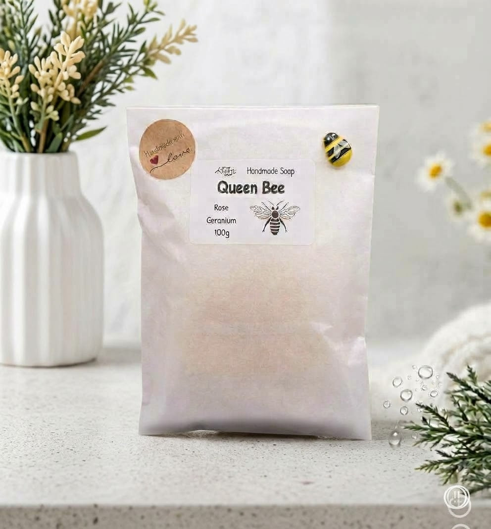 A single package of 'Queen Bee' soap. The off-white paper bag with a clear window revealing the light tan soap is standing upright. The main product label, featuring the title 'Queen Bee', ingredients (Rose, Geranium), and '100g', is visible. A round brown sticker ('Handmade with love') and a small yellow enamel bee pin are attached to the bag. The background is the same well-lit spa environment.