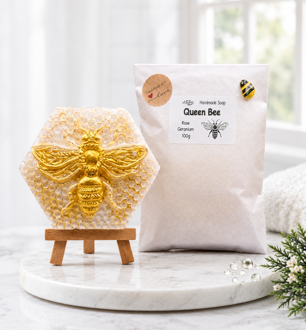A complete set of product images and packaging, presented in a clean, spa-like bathroom. On the left, a hexagonal honeycomb-textured bar of soap with a raised golden bee is displayed on a small wooden easel and a round marble plate. On the right stands the full 'Queen Bee' paper soap package, complete with all stickers and labels, and a small bee enamel pin, showcasing the entire presentation.