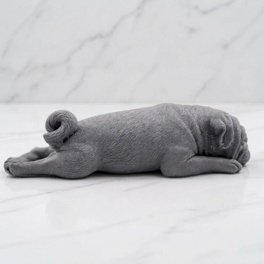 A lying down charcoal grey pug-shaped vegan soap gift. resting on a light marble surface with a soft background.