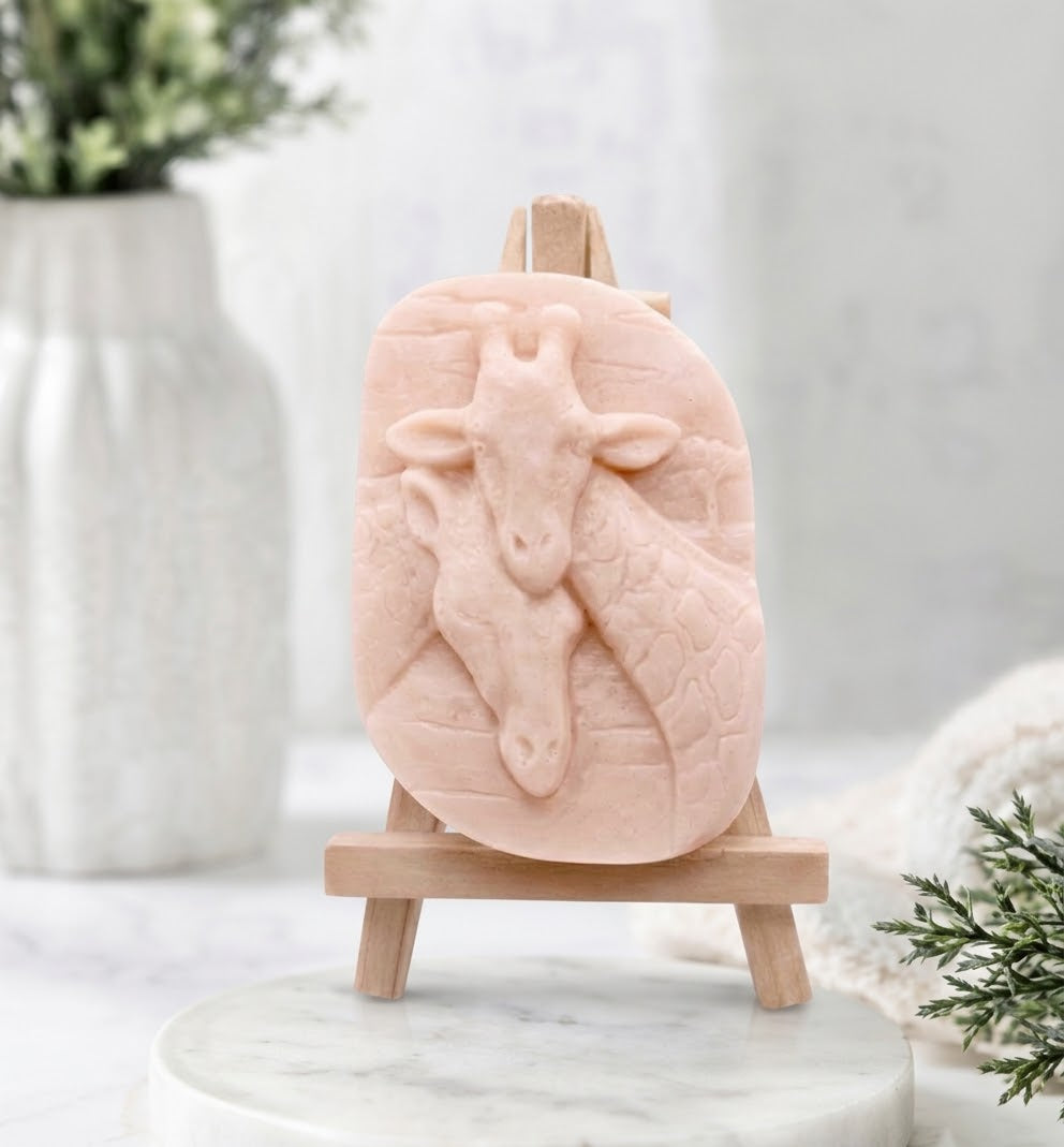 A close-up photograph of a pink coloured handmade vegan soap sculpted into two detailed giraffe heads pressed together in a loving embrace. The soap is displayed on a small, light-wood artist's easel, resting on a white marble surface.