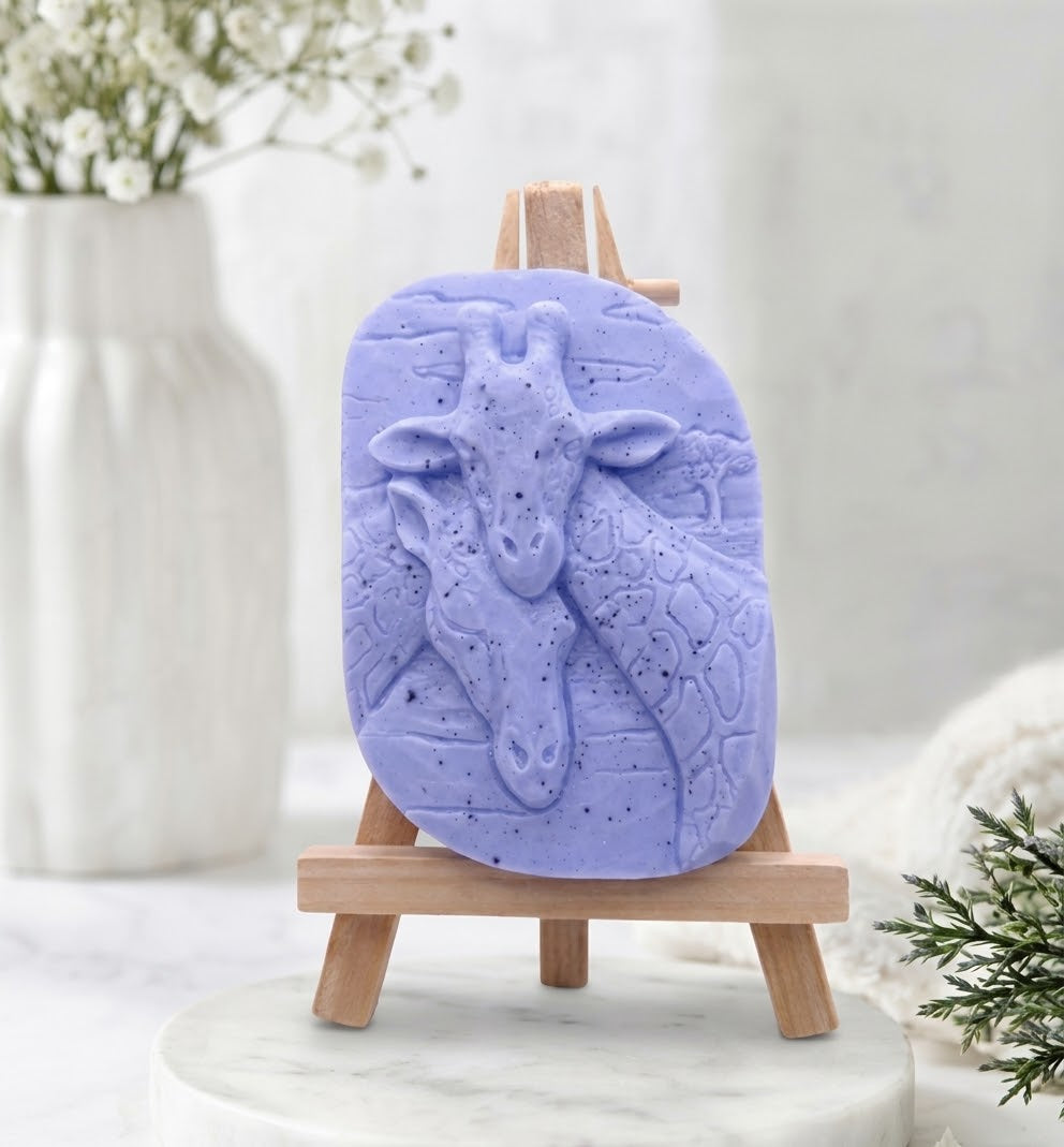 A close-up photograph of a soft blue coloured handmade vegan soap sculpted into two detailed giraffe heads pressed together in a loving embrace, with fine speckles. The soap is displayed on a small, light-wood artist's easel, resting on a white marble surface.