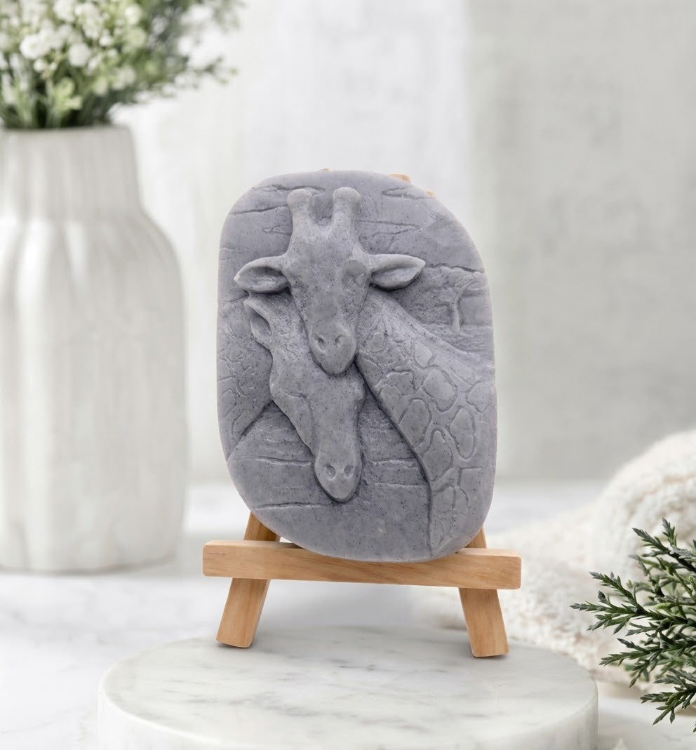 A close-up photograph of a charcoal grey coloured handmade vegan soap sculpted into two detailed giraffe heads pressed together in a loving embrace. The soap is displayed on a small, light-wood artist's easel, resting on a white marble surface.