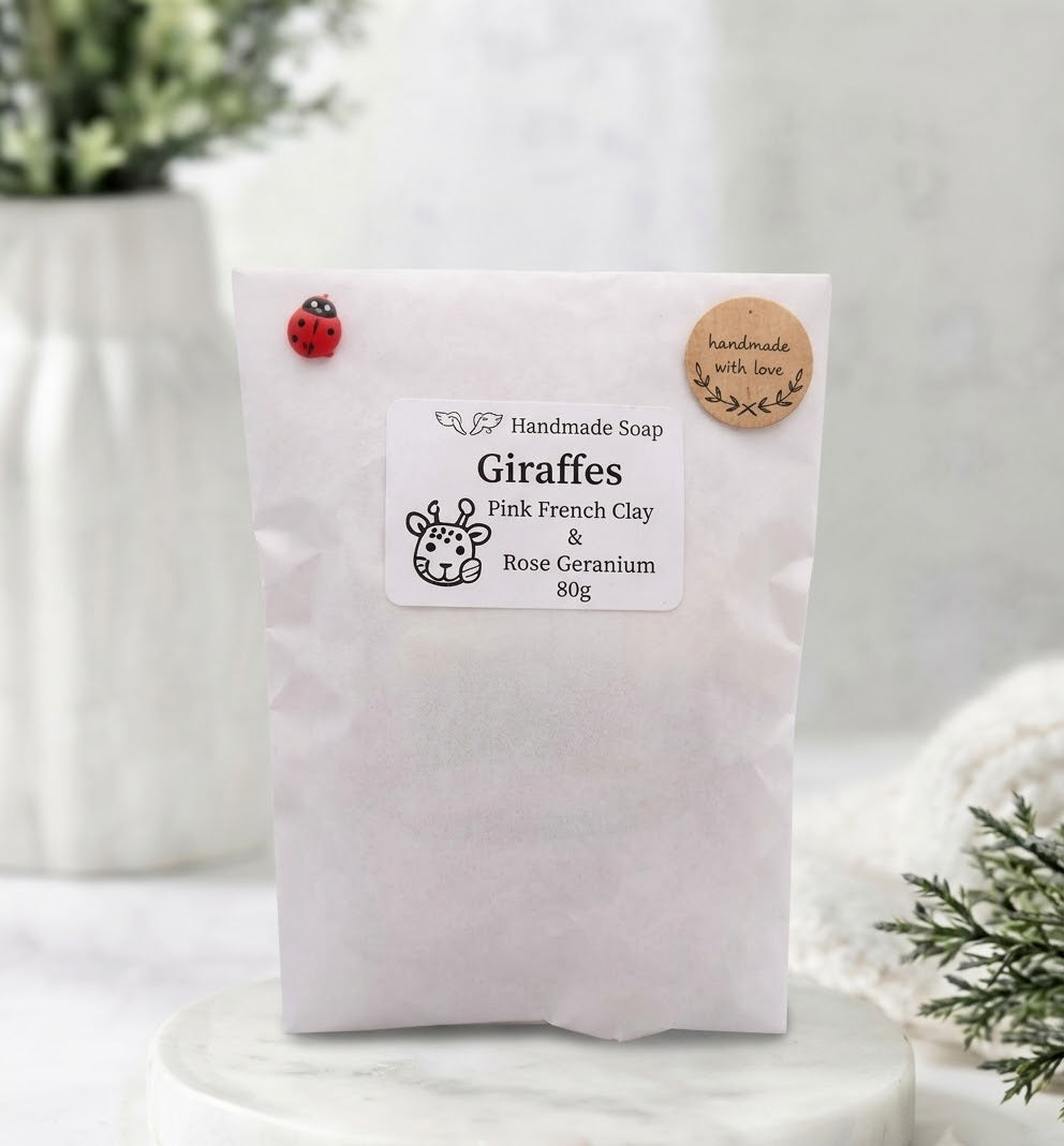 A photograph of the packaging for the "Giraffes" handmade vegan soap, resting on a white marble surface. It is a white paper pouch with a clear window. The label reads: "Handmade Soap Giraffes Pink French Clay & Rose Geranium 80g". A small red ladybug pin is attached at the top-left, and a round "handmade with love" sticker with a heart is at the top-right.