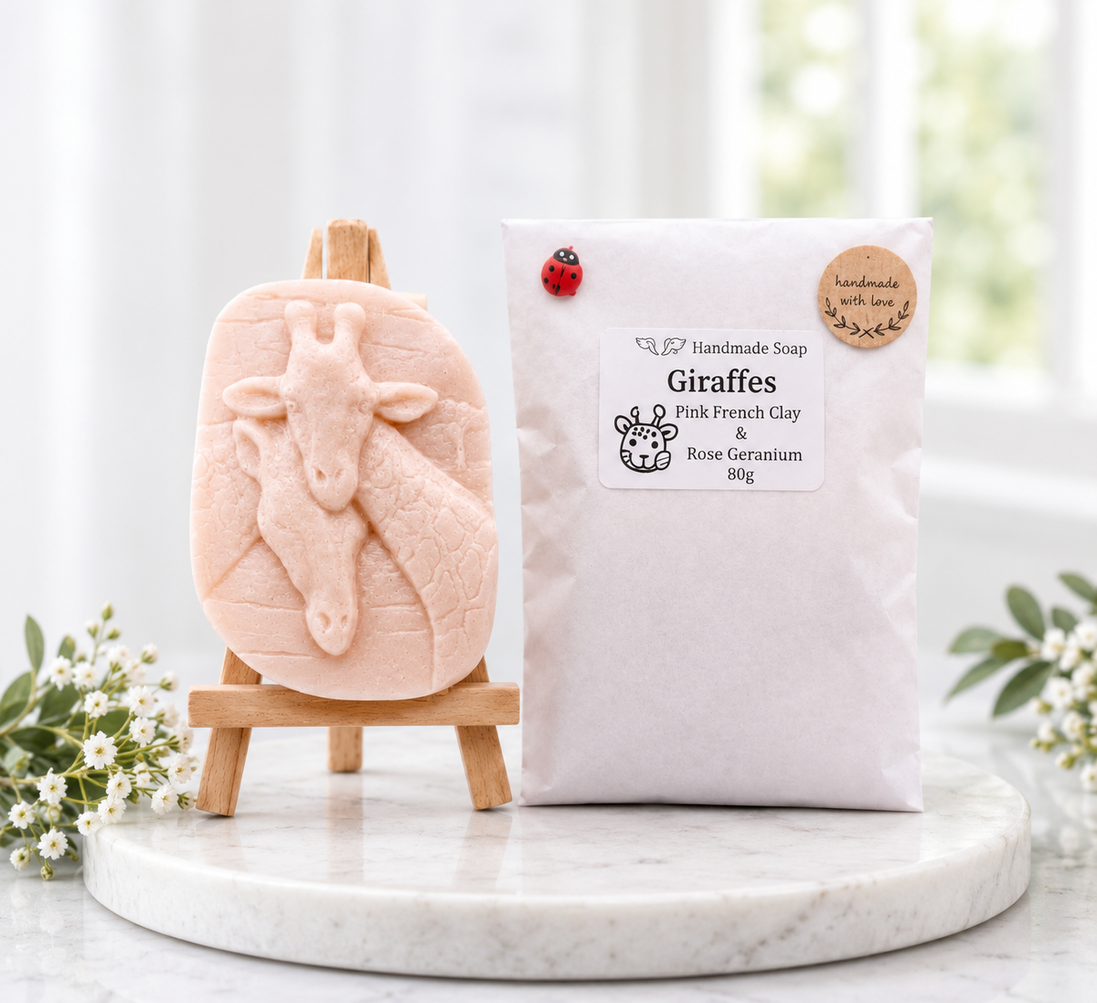 A photograph of a handmade oval soap bar featuring a detailed raised carving of two giraffes resting their heads together. The soap is displayed on a mini wooden artist easel beside its white paper packaging pouch, which features a product label and a small red ladybug accent.