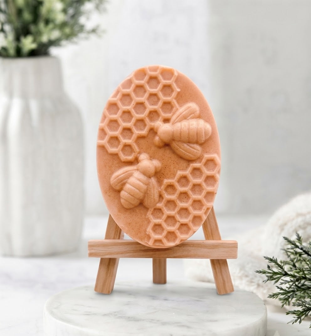 An oval, terracotta-orange coloured soap bar featuring an intricate raised design of two bees and a honeycomb cluster. The soap is standing on a miniature wooden display easel.