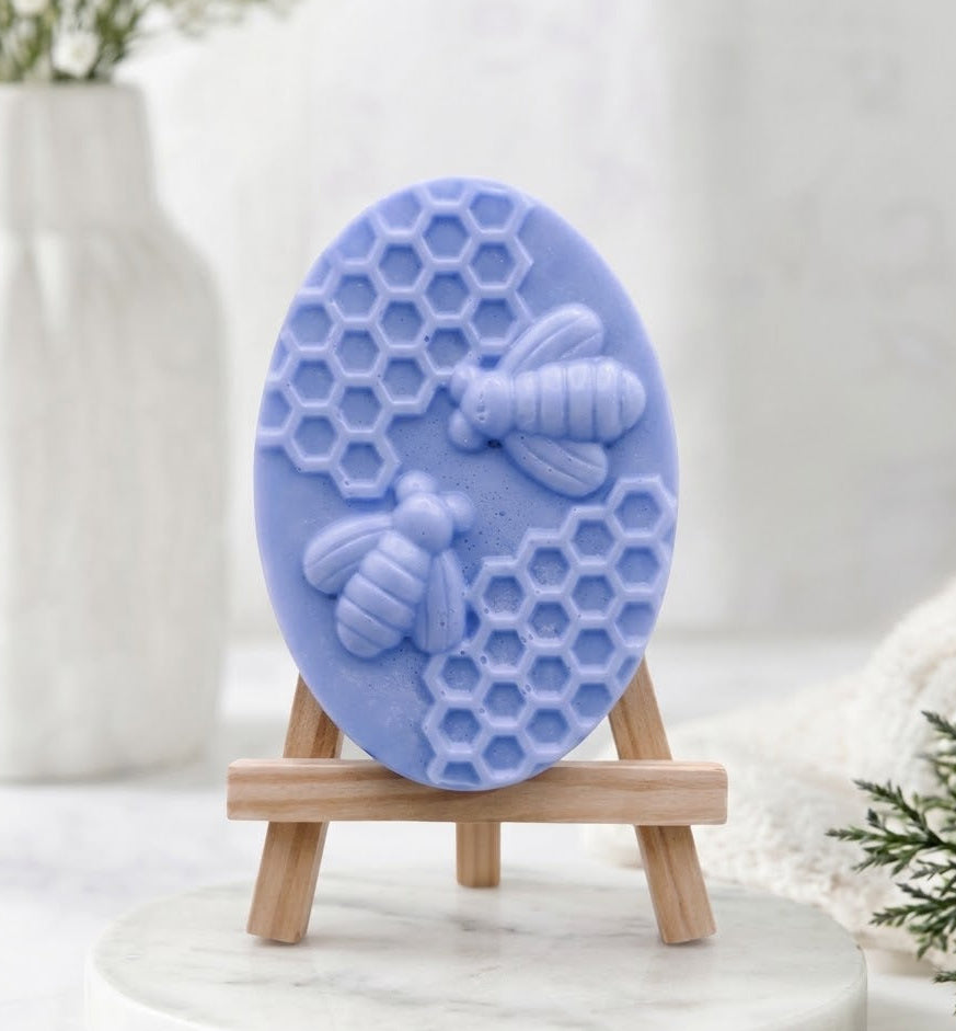 A detailed shot of an oval-shaped, light-blue soap bar featuring a relief carving of two honeybees and a honeycomb pattern. The soap is displayed on a miniature wooden easel.