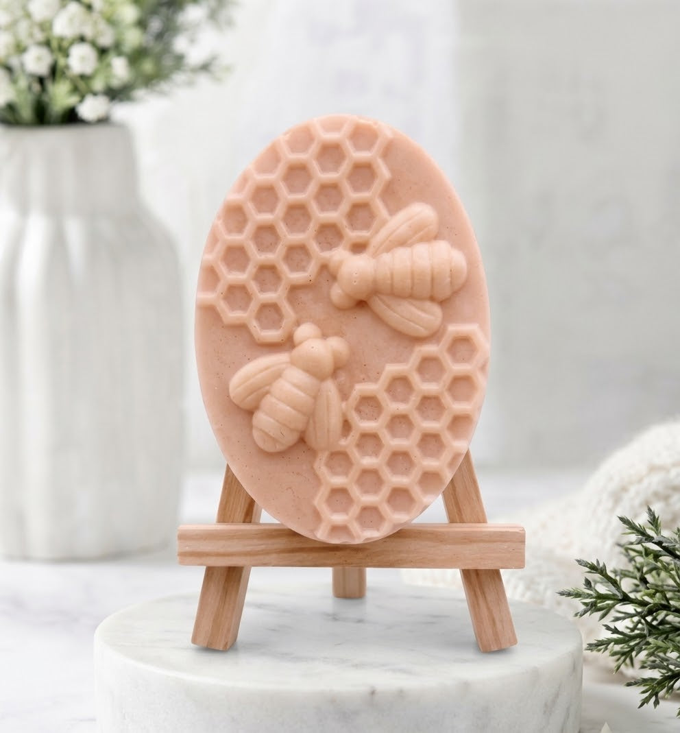 A soft pink coloured oval soap bar with a sculpted relief design of two bees and honeycomb. The soap is displayed on a small wooden artist's easel against a softly blurred background.