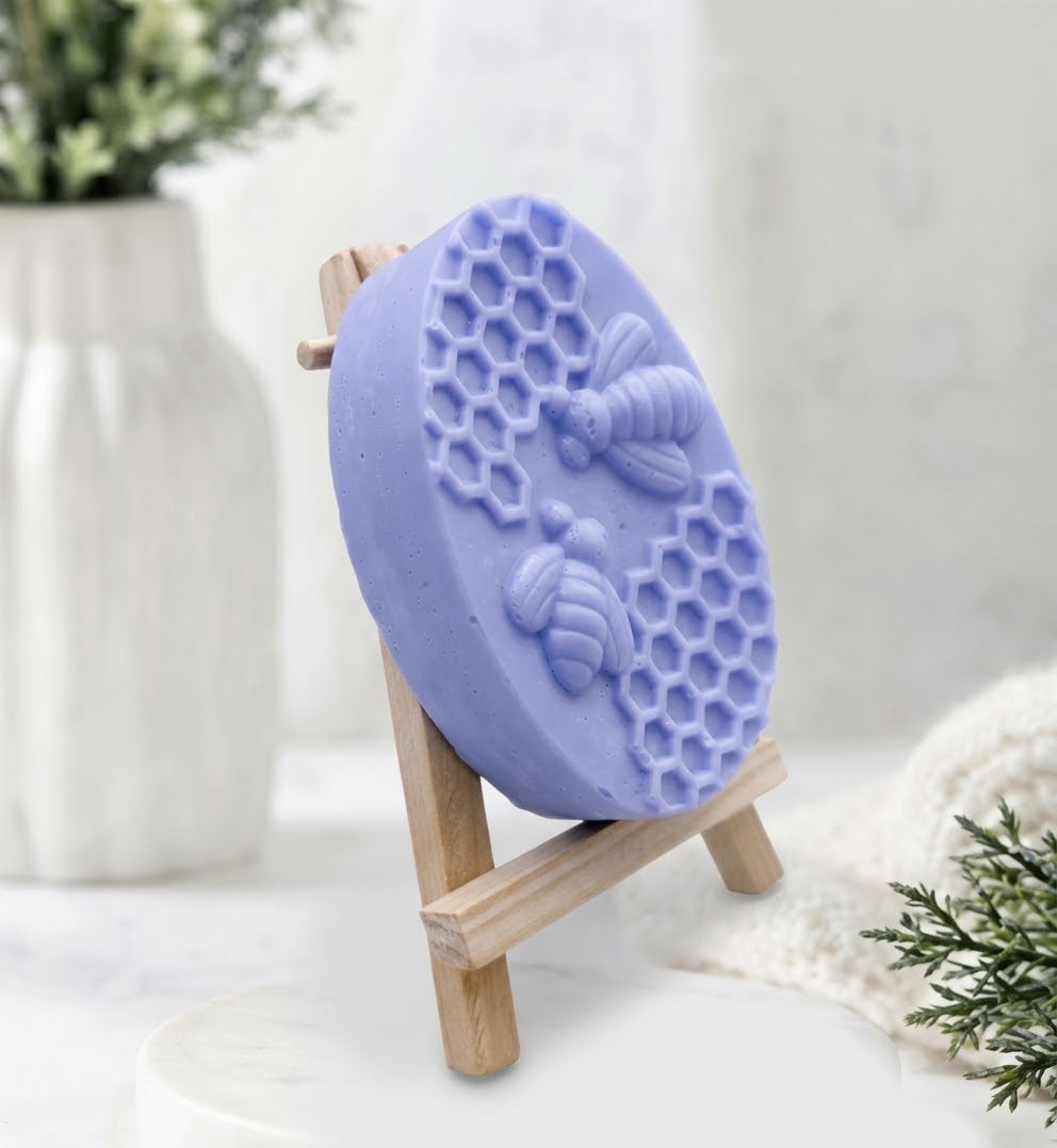 A detailed side view shot of an oval-shaped, light blue soap bar featuring a relief carving of two honeybees and a honeycomb pattern. The soap is displayed on a miniature wooden easel.