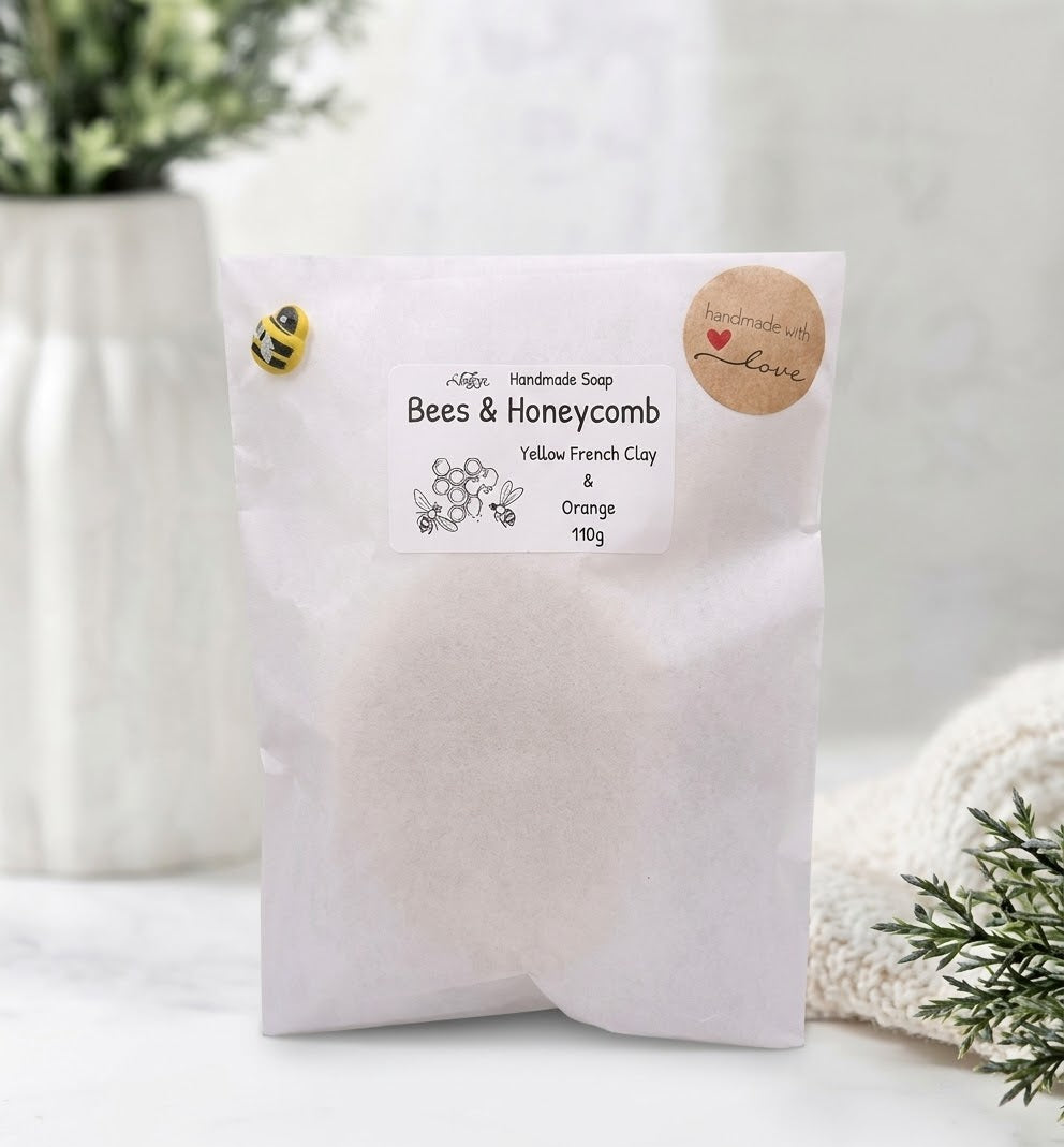 A plain white paper bag package for the handmade bee soaps. It is sealed with a round kraft paper sticker that says "handmade with love" and features a small, decorative yellow and black bee charm pinned to the top left. The central label reads "Handmade Soap: Bees & Honeycomb - Yellow French Clay & Orange - 110g."
