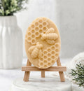 A soft yellow-toned, beige oval soap bar with a sculpted relief design of two bees and honeycomb. The soap is displayed on a small wooden artist's easel against a softly blurred background.