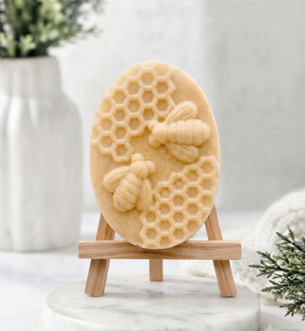 A soft yellow-toned, beige oval soap bar with a sculpted relief design of two bees and honeycomb. The soap is displayed on a small wooden artist's easel against a softly blurred background.