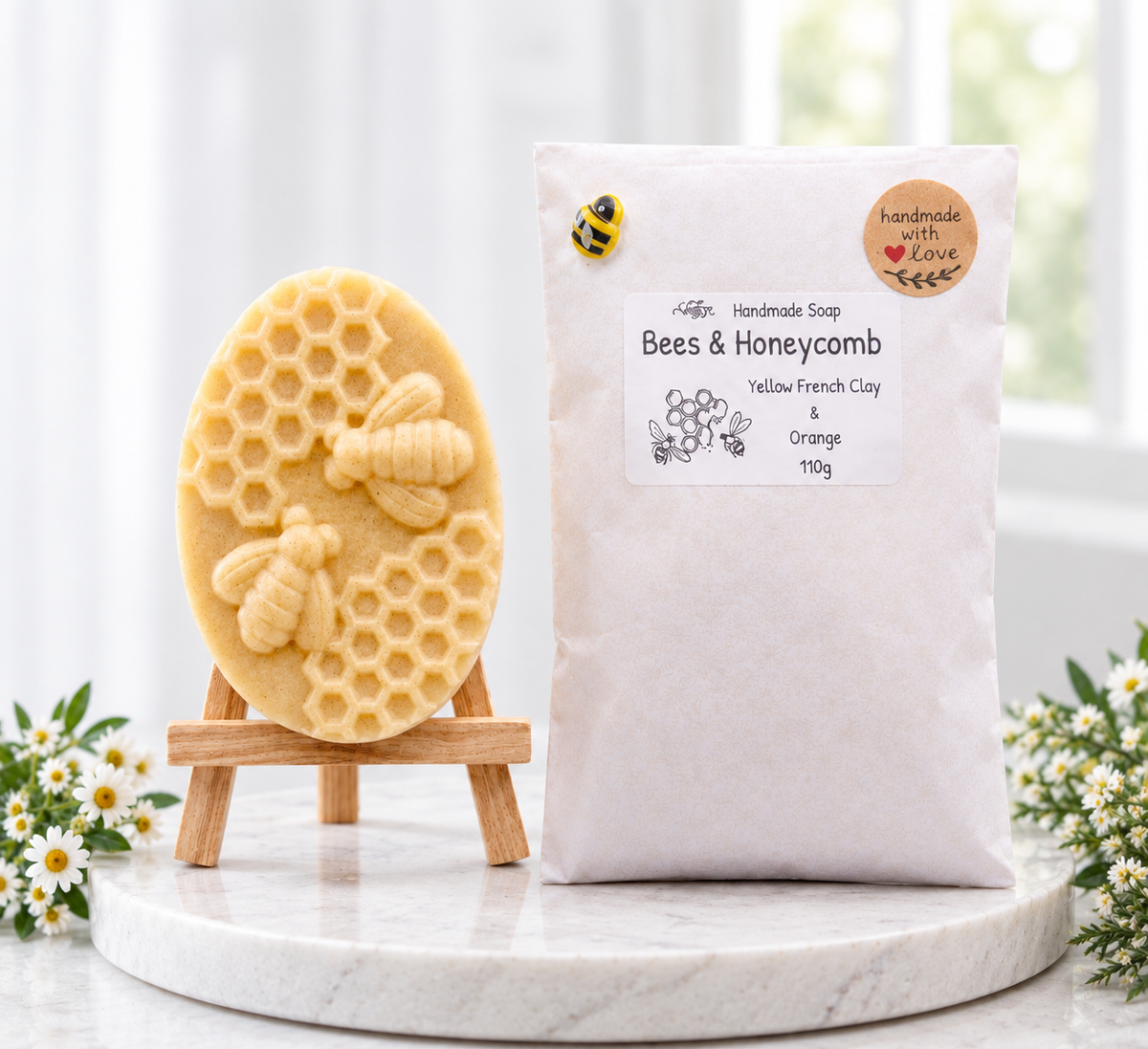 A photograph of a handmade oval yellow-cream soap bar featuring a detailed raised carving of two bees and a honeycomb pattern, displayed on a small wooden easel. Beside it sits the white paper packaging pouch with a product label reading "Bees & Honeycomb", a small bumblebee embellishment, and a "handmade with love" sticker. Both items are set on a round white marble display slab against a bright, softly blurred background framed with white flowers.