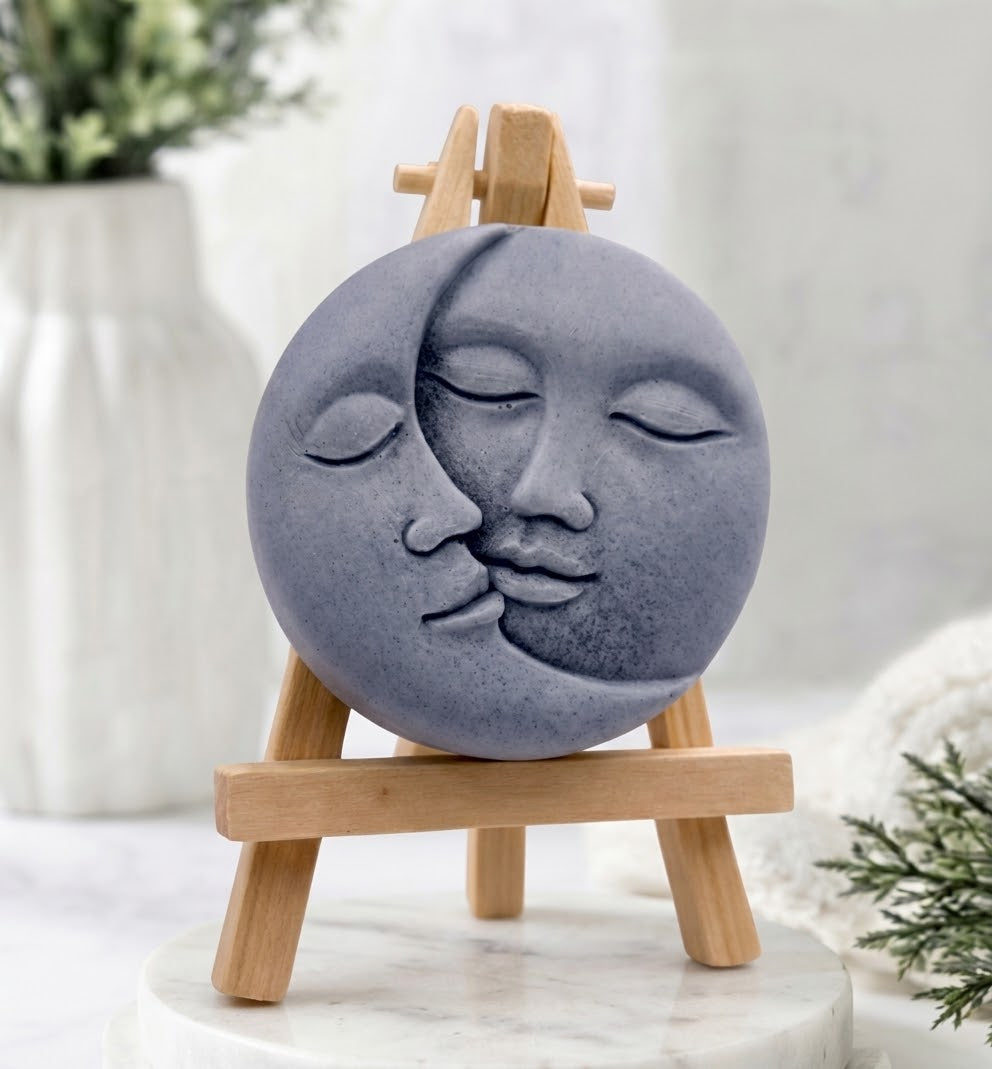 A grey, round vegan soap with a carved sun and moon kissing face design.