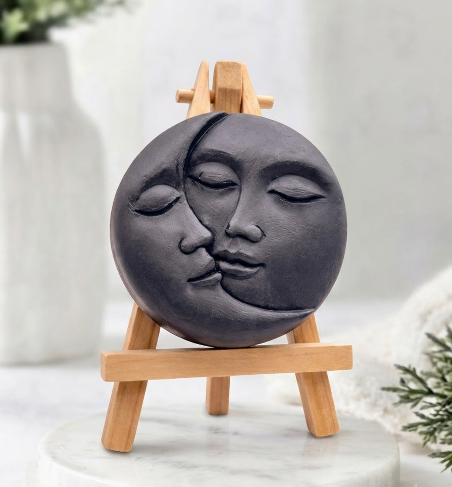 A charcoal Jet black, round vegan soap featuring a sun and moon kissing face carving.