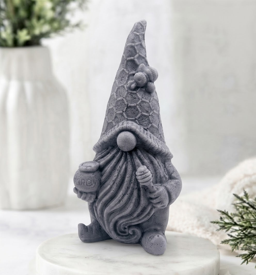 A textured dark grey gnome-shaped vegan soap, featuring a detailed beard, honeycomb-patterned hat, and bee decoration. The background shows a white ribbed vase with leaves and a knitted towel.