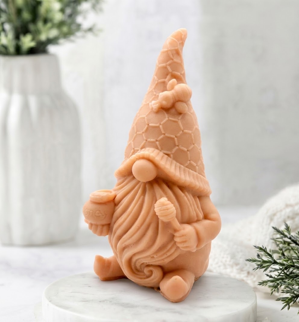 A direct close-up photograph of a detailed terracotta coloured gnome-shaped vegan soap on a white marble pedestal. The intricate details include a detailed beard, honeycomb hat, and a tiny bee decoration. The background features a full view of a white vase with foliage.