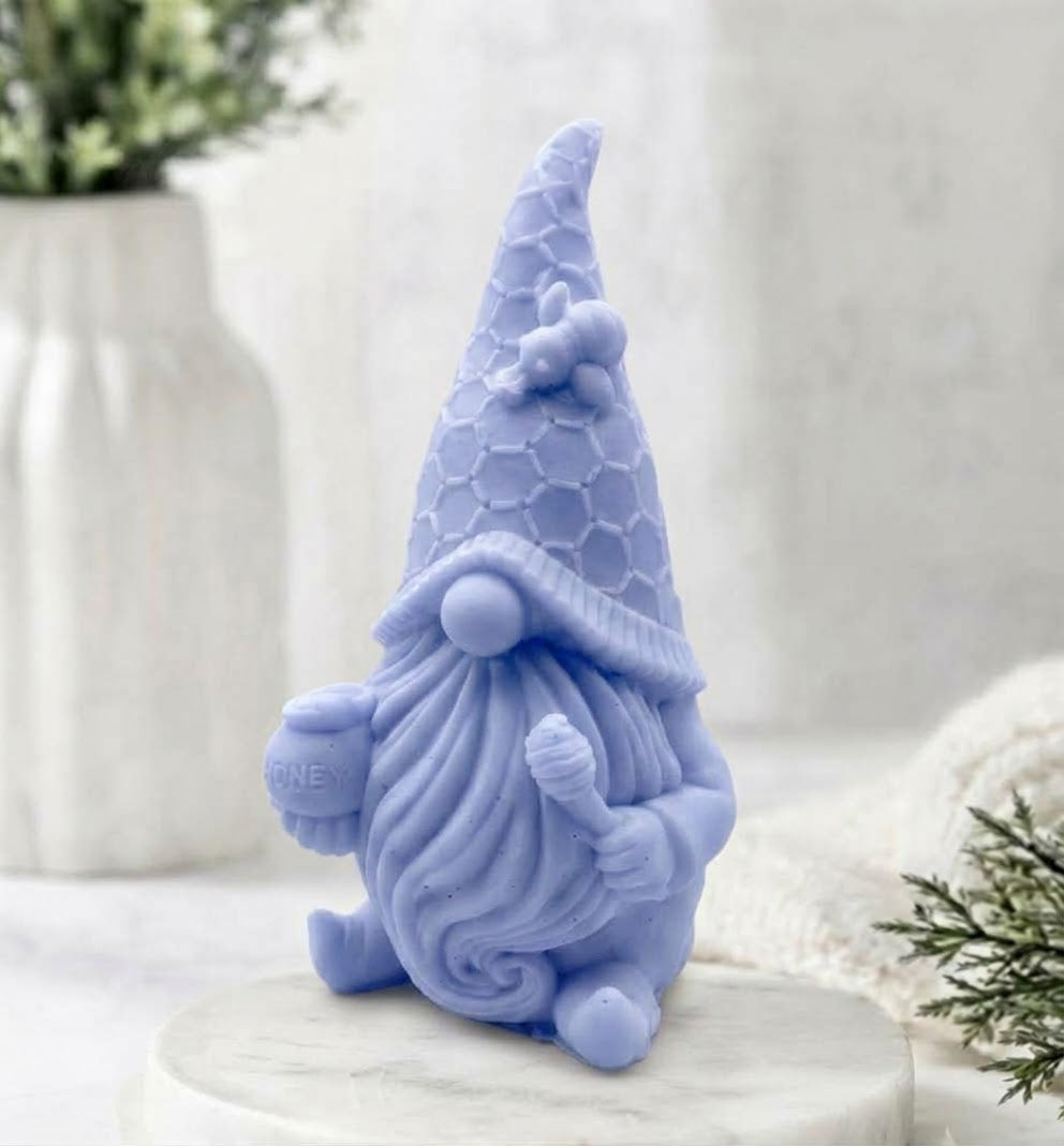 A detailed close-up shot of a distinct light blue gnome-shaped vegan soap on a white marble coaster, showcasing its detailed beard, honeycomb hat, and bee decoration. The background includes a blurred vase and white knit fabric.