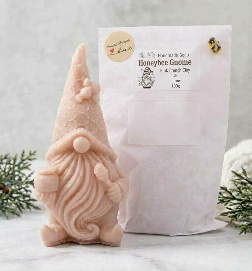 A direct view of a detailed pink-clay gnome-shaped vegan soap beside its white paper packaging bag. The bag features a main label reading "Honeybee Gnome Pink French Clay & Lime 100g" and a round "Handmade with Love" sticker.