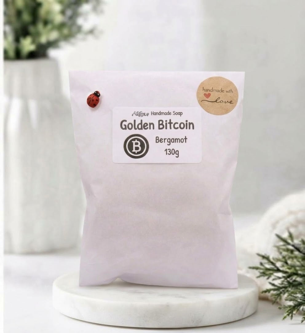 A photograph of the 'Golden Bitcoin' vegan soap, presented in a clean, white paper bag packaging. A small, red and black ladybug pin is clipped to the upper left corner, and a brown paper sticker on the upper right reads, "Handmade with love". The main product label in the centre displays a black Bitcoin symbol and text