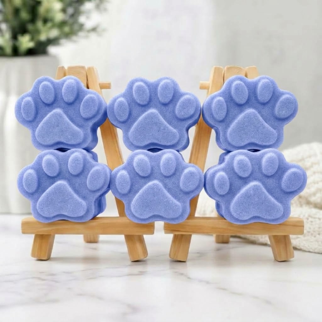 A collection of six blue paw print-shaped soaps, arranged in three stacks of two on a wooden easel. Greenery and a blanket are in the background.