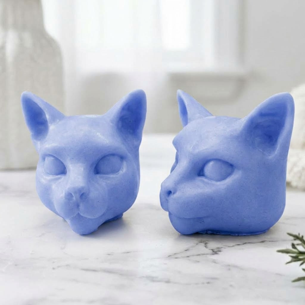 Two detailed, light lavender-blue vegan soap bars handmade into accurate Siamese cat heads, resting on a white marble surface against a blurry bathroom background.