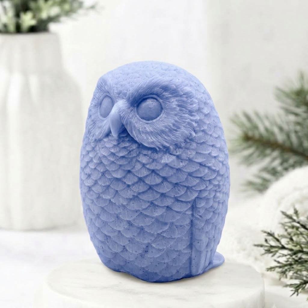 A detailed indigo blue handmade vegan soap in the shape of an owl with intricate feather textures, displayed on a light wooden table with white flowers in the background.