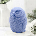 A detailed indigo blue handmade vegan soap in the shape of an owl with intricate feather textures, displayed on a light wooden table with white flowers in the background.