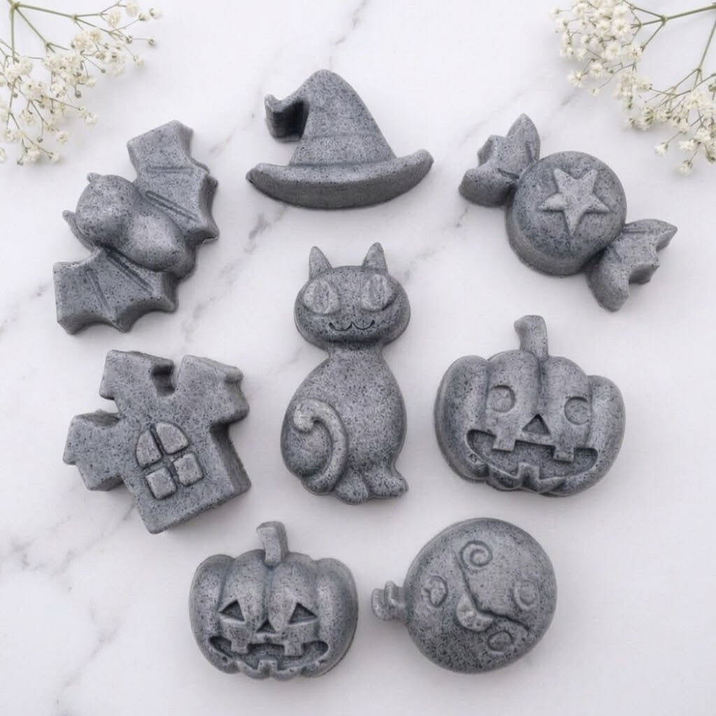 A charcoal grey collection of eight handmade vegan Halloween soaps featuring a bat, witch's hat, star-patterned sweet, haunted house, black cat, jack-o'-lantern, and a ghost, arranged on a light wooden surface with white floral accents.